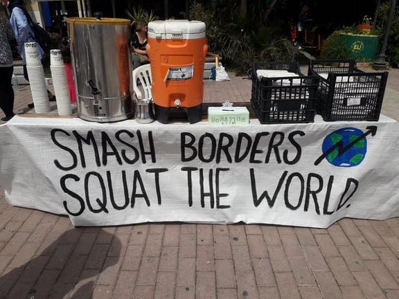 A picture of a food stand with tea, coffee and donation box. And a banner that says: SMASH BORDERS, SQUAT THE WORLD